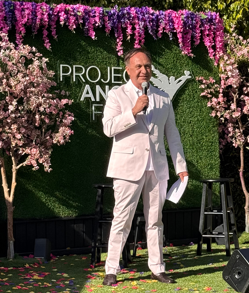 Asm. Zbur holding microphone and speaking, with foliage backdrop in background