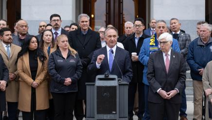 Asm. Zbur at podium, speaking, with multiple officials standing in support