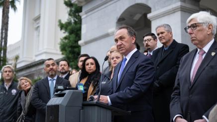 Asm. Zbur at podium, speaking, with multiple officials standing in support