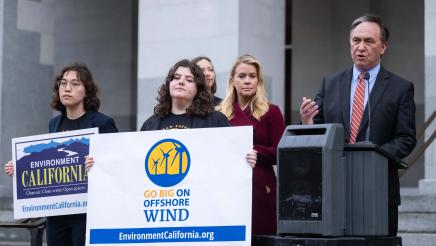 Asm. Zbur at podium, speaking, with two participants holding up large signs